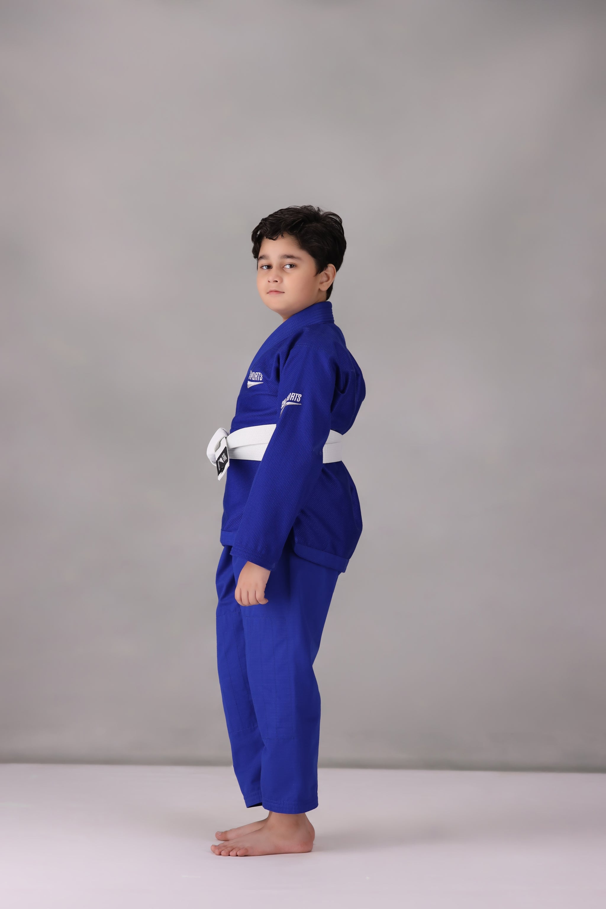 Child wearing a custom kids Jiu Jitsu Gi in the UK, blue Brazilian Jiu Jitsu uniform with a white belt on a gray background