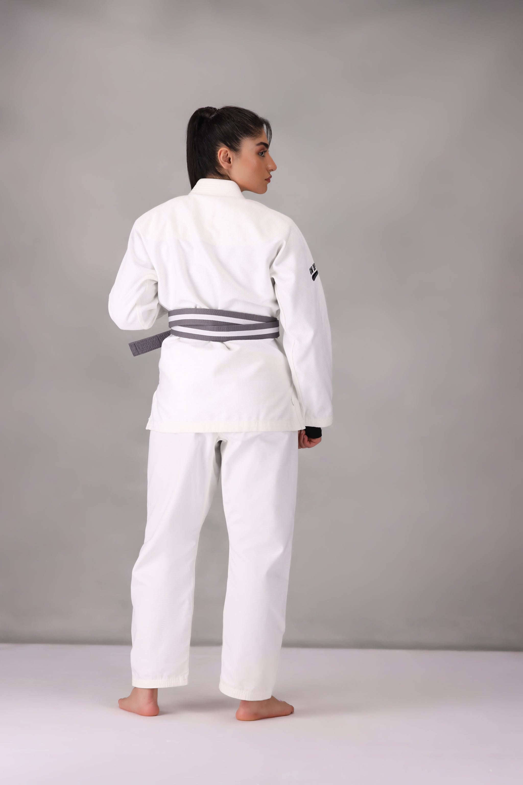 Woman wearing a custom white BJJ Gi with a black belt, standing on a gray background UK Brazilian Jiu Jitsu uniform