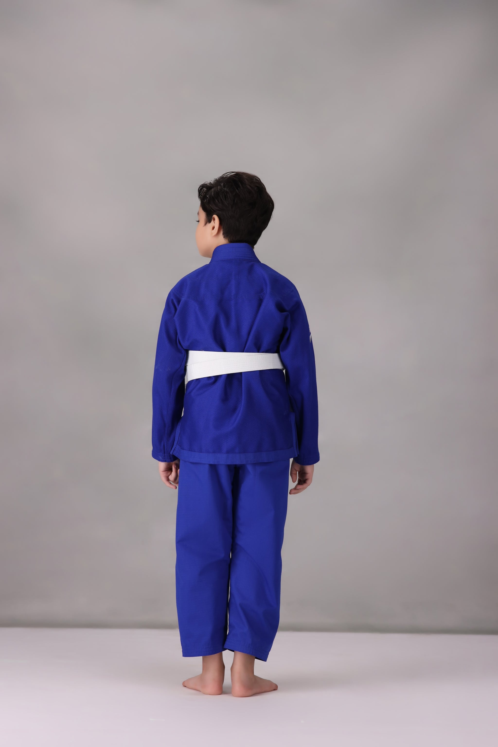 Child wearing a custom kids Jiu Jitsu Gi in the UK, blue Brazilian Jiu Jitsu uniform with a white belt on a gray background