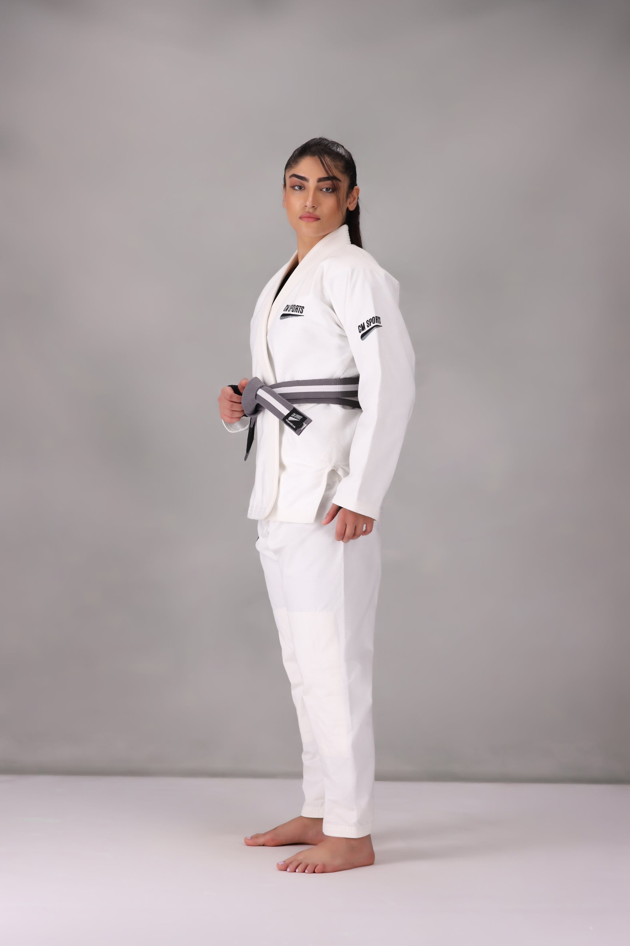 Woman wearing a custom white BJJ Gi with a black belt, standing on a gray background UK Brazilian Jiu Jitsu uniform